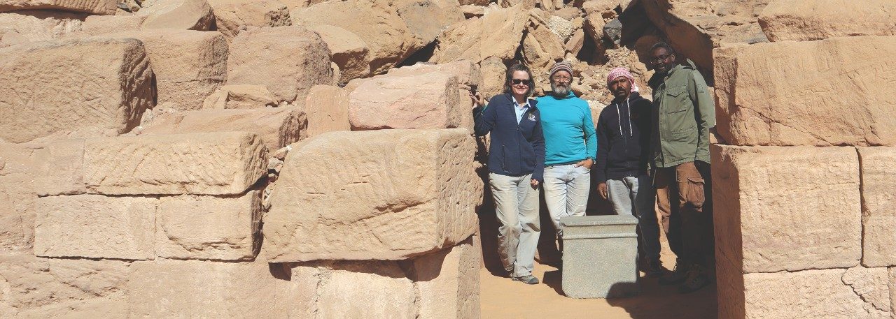 A group of four people stands in the remains of a temple consisting of large stacked sand-stone blocks covered with shallow inscriptions.