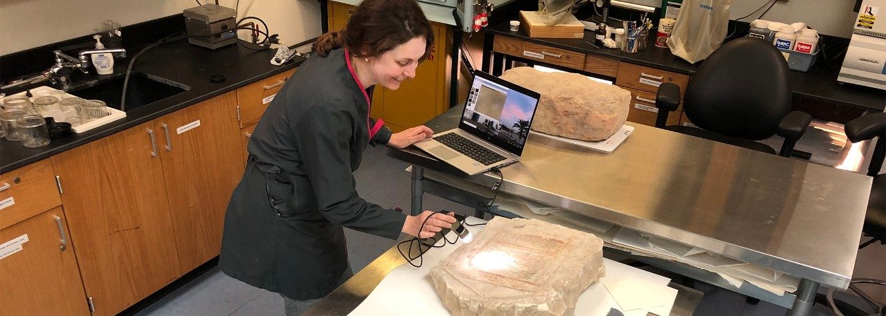 Conservator Carrie Roberts holds a small digital microscope with a light over a limestone grave marker to view pigments on its surface. Near her on a table are two other large stelae.