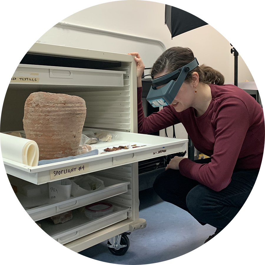 Carrie Roberts, wearing a headband magnifier, squats down to closely examine objects within the drawer of a storage cabinet. The drawer is labeled with the words “Spotlight #4, and visible artifacts include a terracotta vessel and textile fragments.