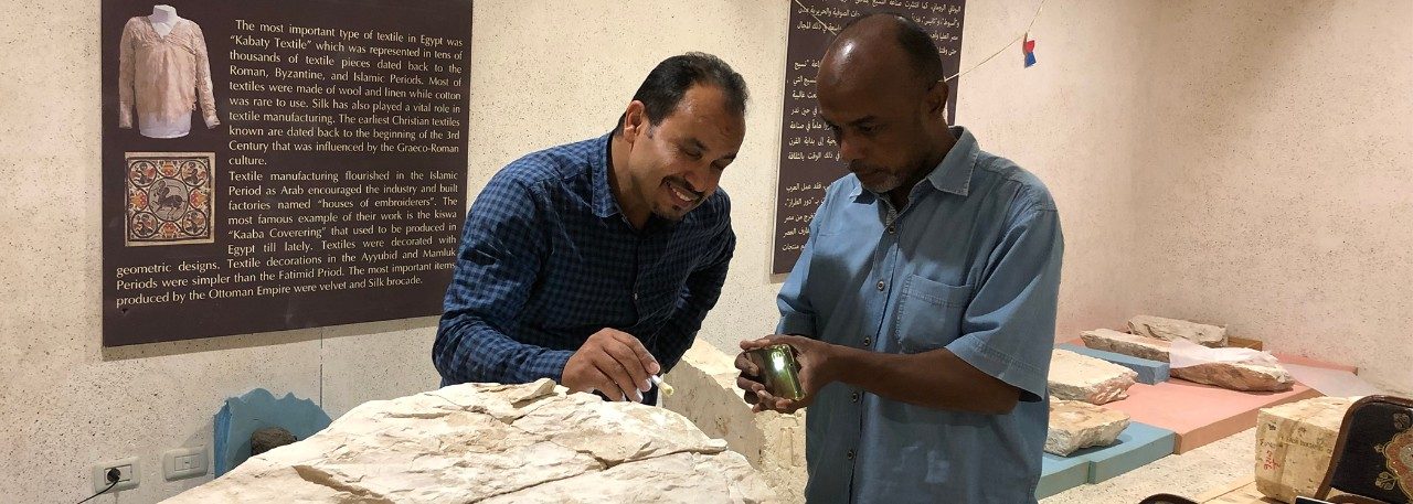 Two men, Hamada Sadek and Ahmed Abdollah, stand in a room of the Sohag National Museum working on a massive stone block. One man shines a Ahmed shines a light from a phone while Hamada uses a small brush on the surface of the object.