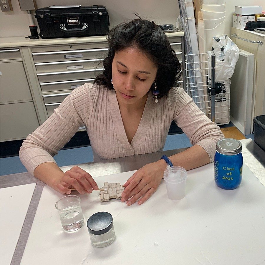Kathryn Peneyra, eyes downcast, wipes a white mosaic fragment with a cotton swab. In front of her are containers filled with various solutions, and to her left is a cart holding a larger mosaic fragment.