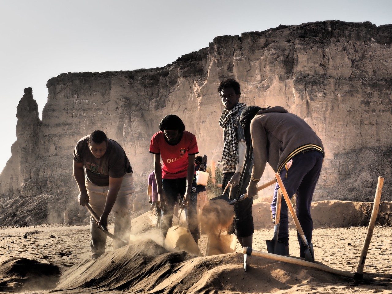 Archaeological work in Sudan. Two men dig with shovels in a sand pile, while two more hold bags open. The large mesa of Jebel Barkal rises In the background.