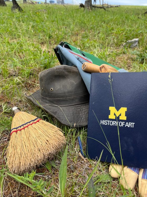 A hat, gloves, broom, and notebook with the logo for the University of Michigan's History of Art Department lying in the grass at an excavation site.