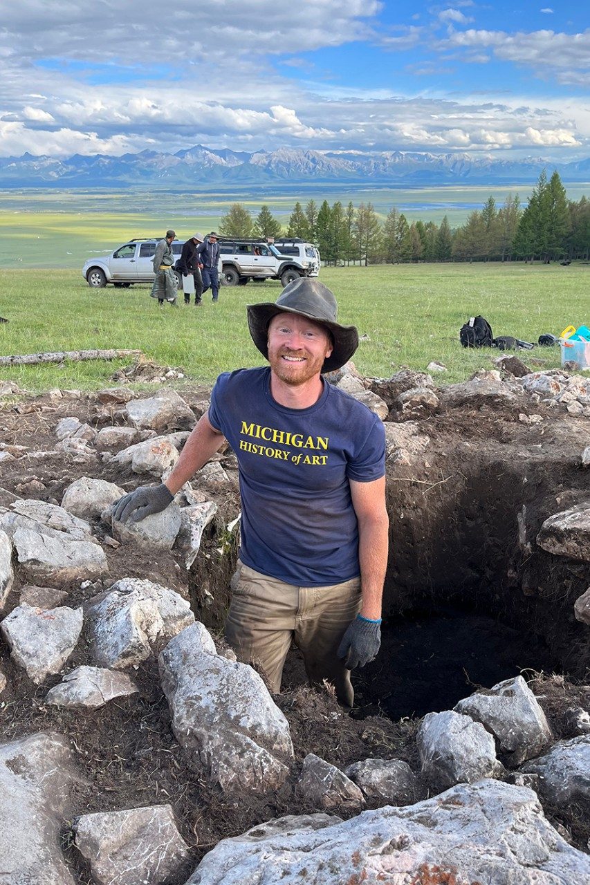 Bryan Miller, wearing a University of Michigan History of Art Department shirt and a hat, standing in a rocky pit at an excavation site. Behind him are vehicles and a small group of people, while a lush landscape of greener, lakes, and mountains is in the background.