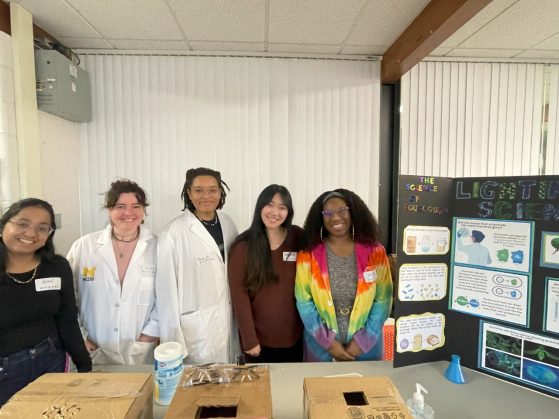 Five women science graduate students and a poster about glow-in-the-dark-bacteria