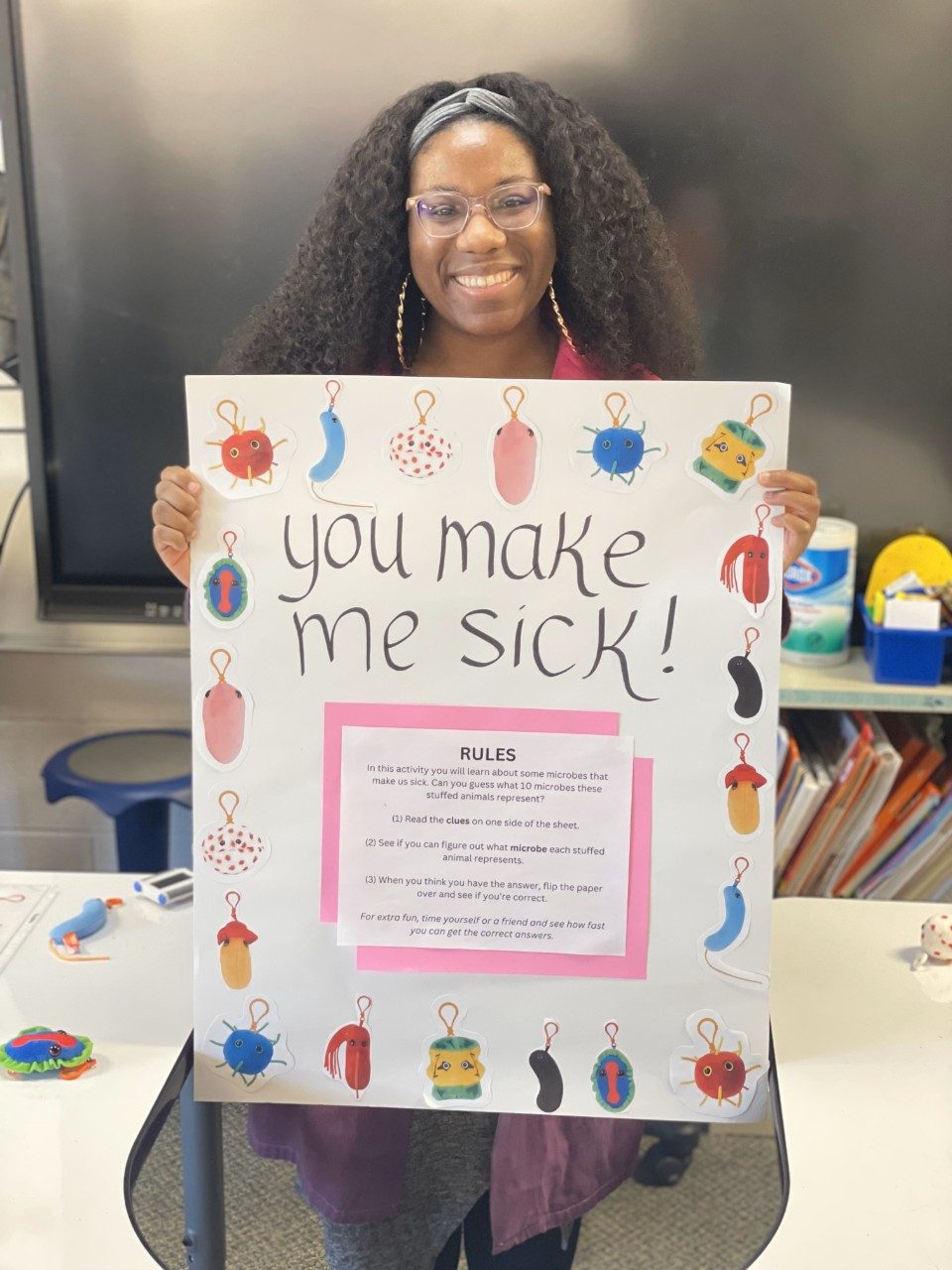 A smiling woman holds up a sign for a game called "you make me sick" 