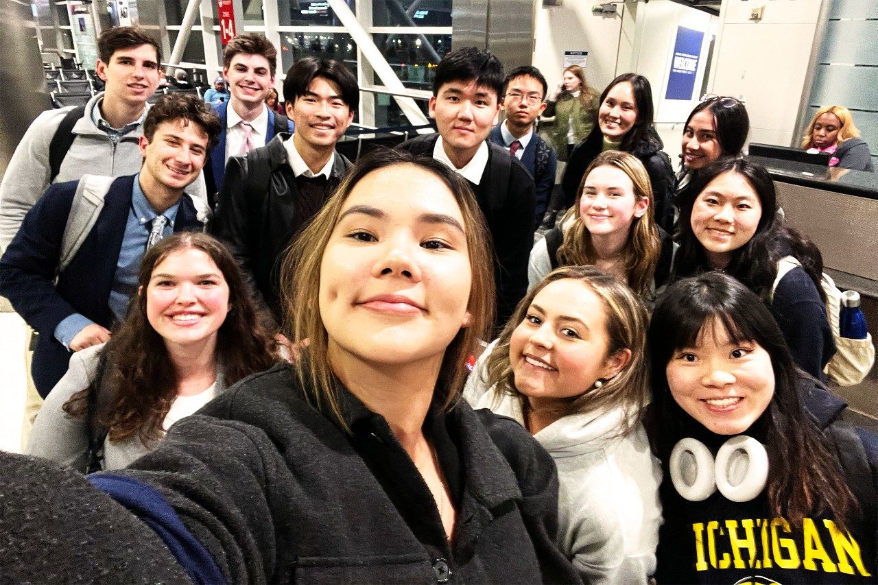 Group of smiling students taking a picture of themselves in selfie mode, some wearing Michigan gear.