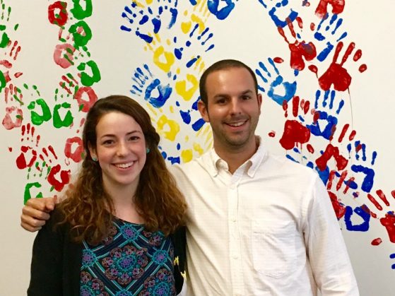 two people smiling in front of a wall covered in colorful hand prints