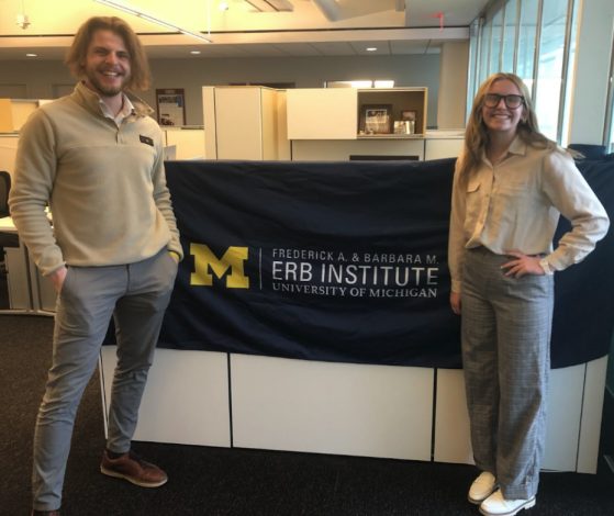 Two people smile together in front of a michigan flag