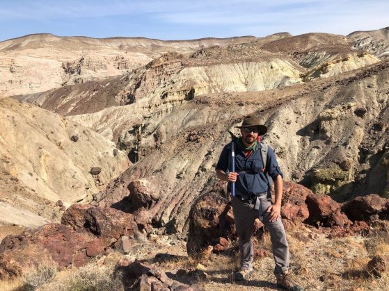 Man stands in wide brimmed hat, holding pick axe over shoulder on top of a rocky mountain