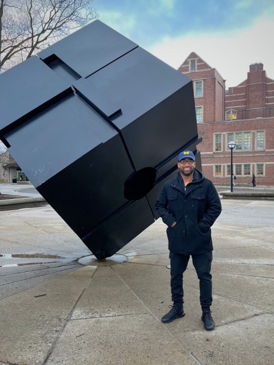 Man standing next to black cube sculpture on U-M campus
