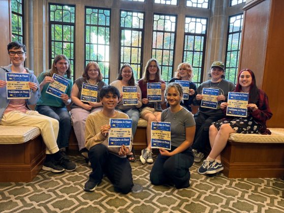 a group of students sit in a lounge in front of leaded glass windows smiling at the camera