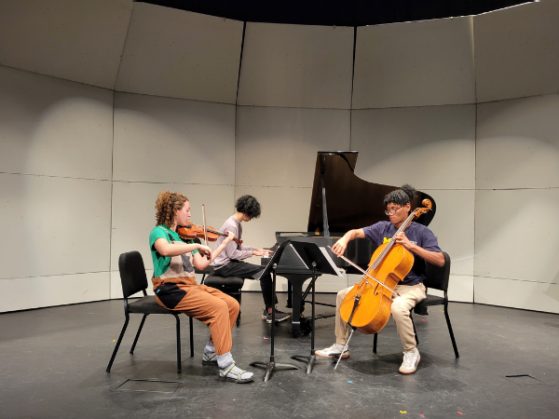 three people perform with instruments on stage including a piano, violin, and cello