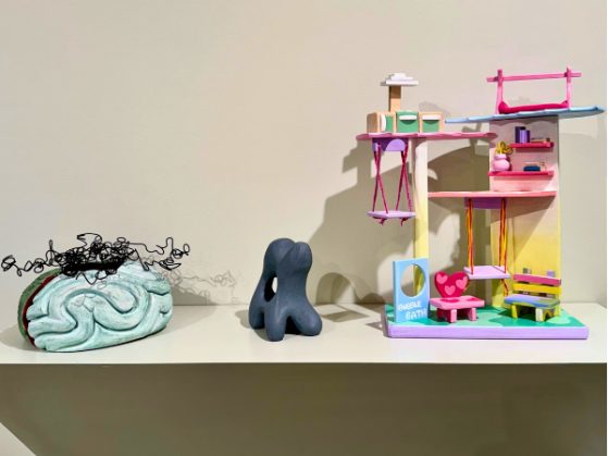 a gallery exhibit featuring a brain-like sculpture with dead flowers growing from the top, a small amorphous body sculpture, and a brightly colored child's play house made from found objects