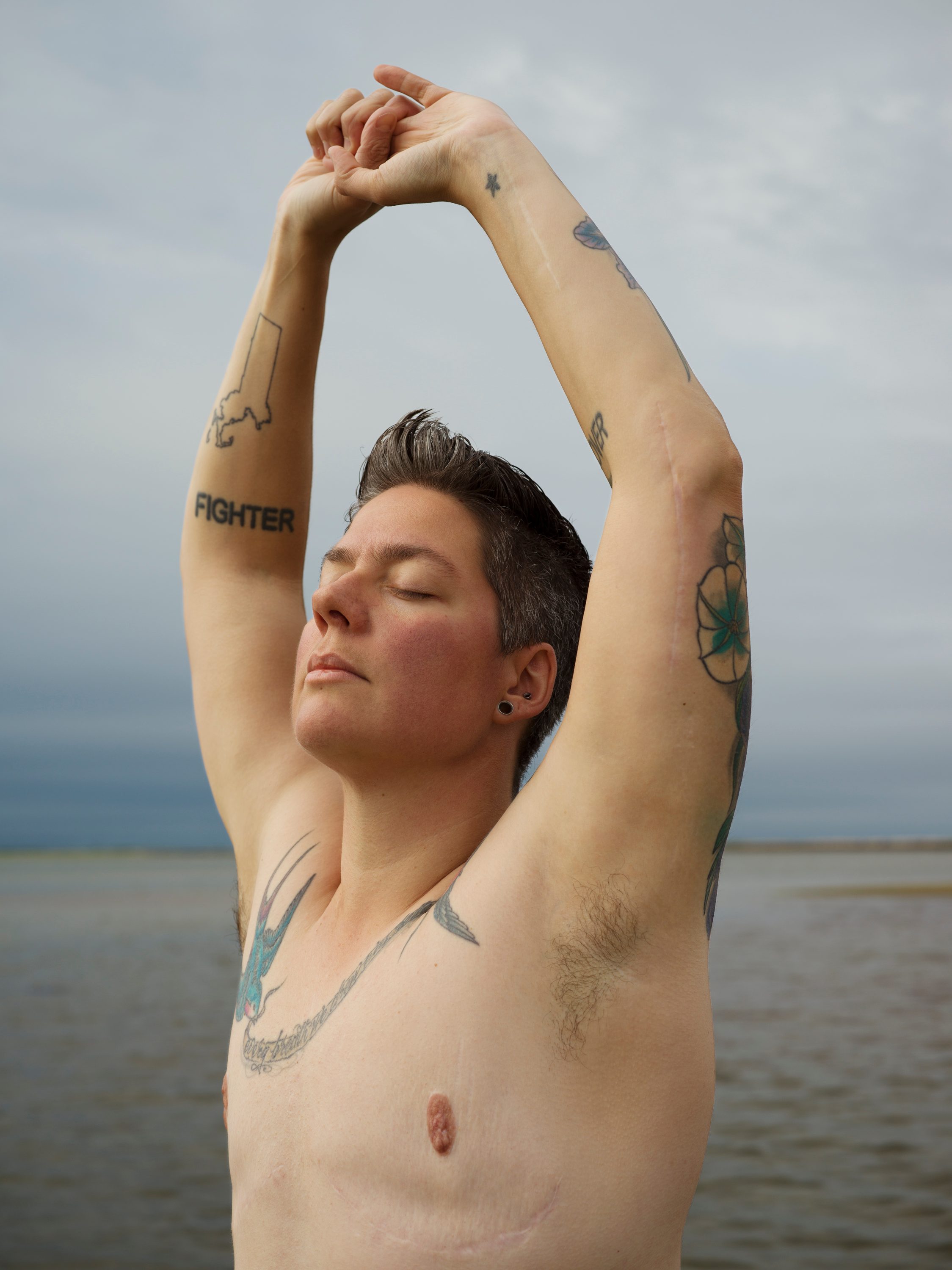 A person stands shirtless, eyes closed with hands stretched over the head. The person looks at peace.