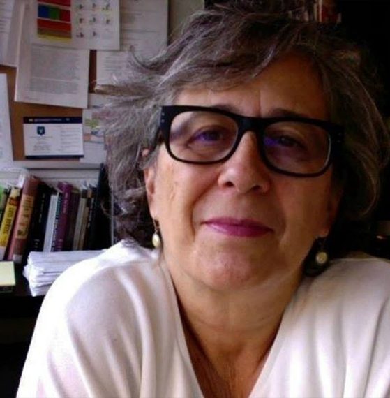 Image of RLL faculty member, Cristina Moreiras-Menor (Spanish Program). A casual photo where she is wearing a white blouse, black frame glasses, and button drop earrings. There is a corkboard with pinned papers and a desk with books on it behind her.