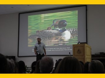 Person on a stage talking in front of an image of loons
