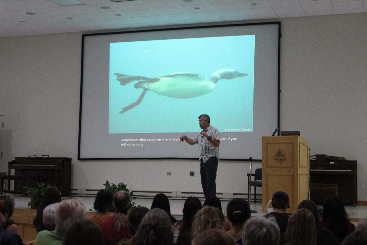 Person talking on stage with a loon on the screen in the background