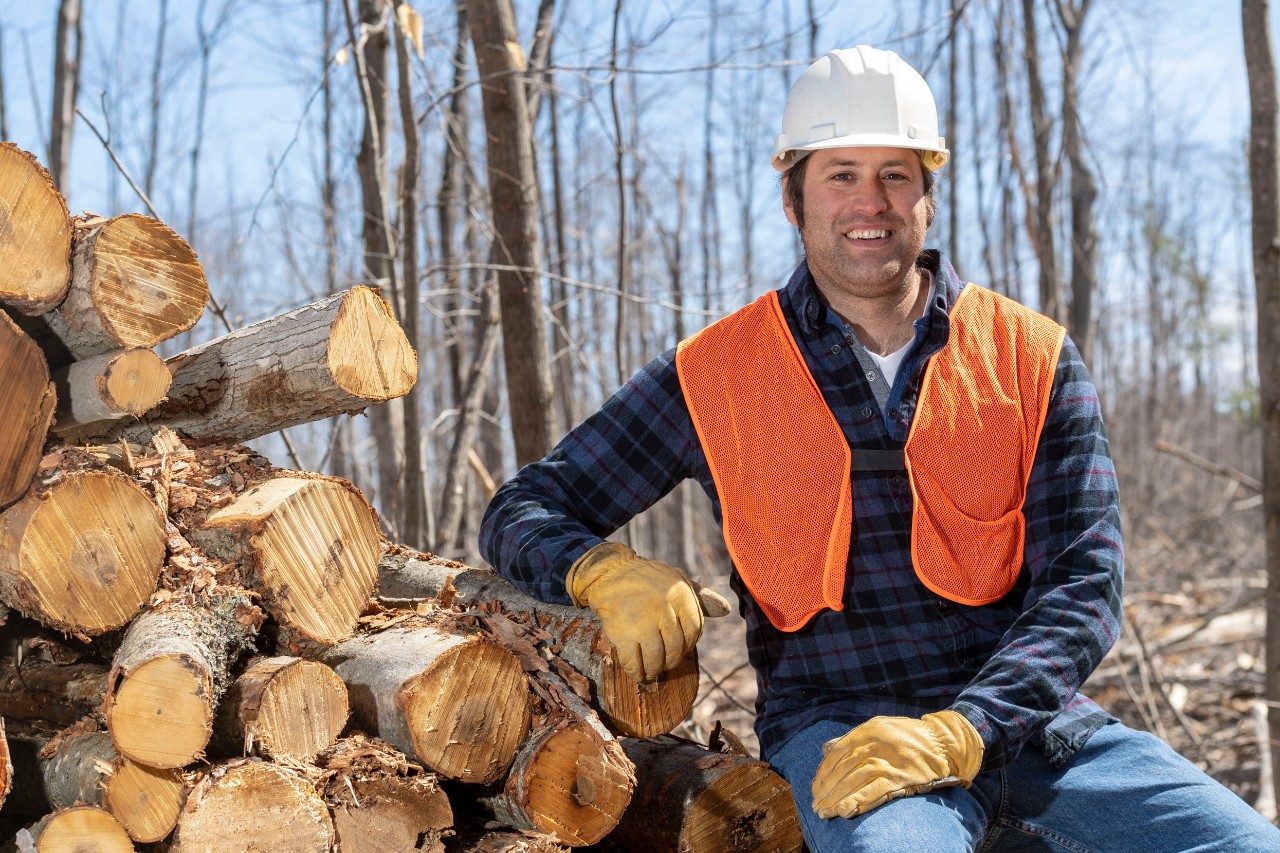 Man wearing a hard-hat and orange vest next to pile of logged trees in a forest
