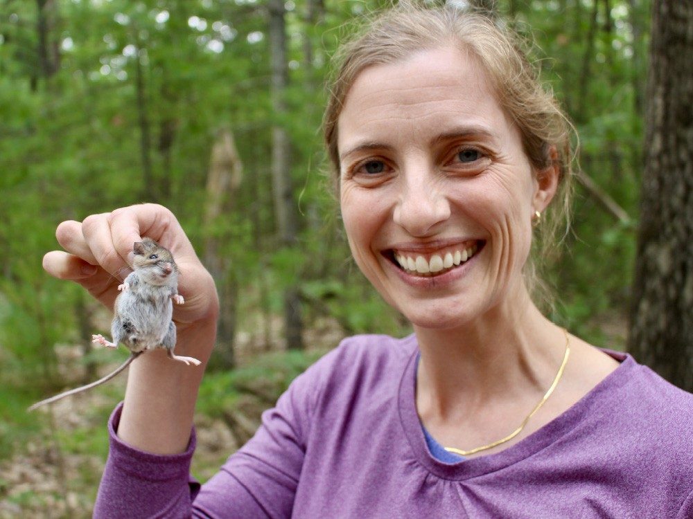 Woman holds mouse by the scruff of its neck in a forest