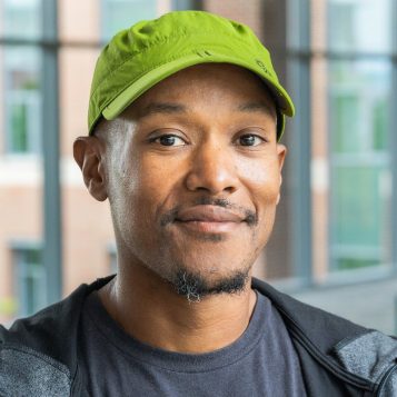 Headshot photo of a man wearing a green hat in front of windows