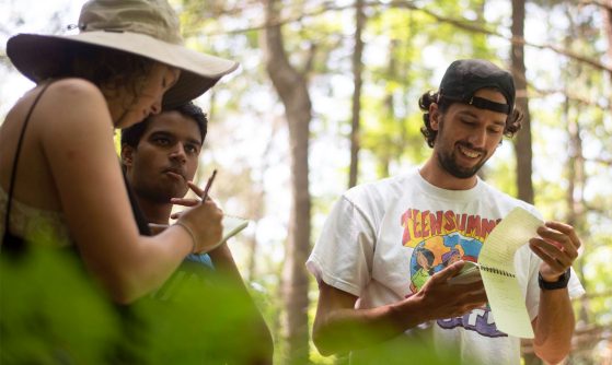 3 students look at note pads in a forest